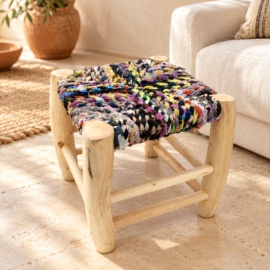 Taburete Tadwir de madera y retales multicolor junto a un sofá en un salón cálido.