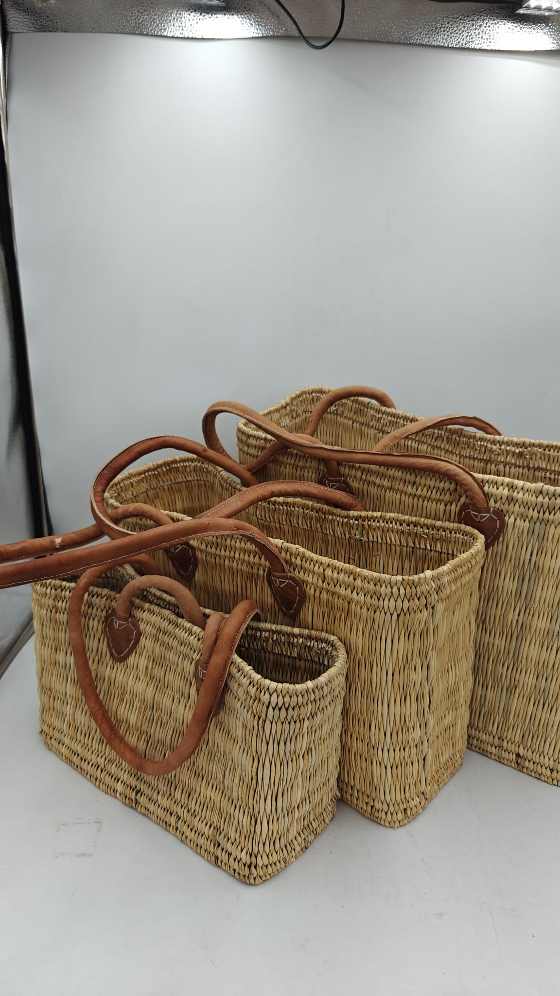 Conjunto de capazos artesanales marroquíes de esparto natural apilados, con asas largas de cuero, vista lateral mostrando los tres tamaños